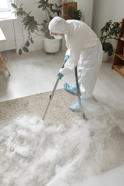 A professional cleaner from Swiss Cottage Carpet Cleaners is performing deep carpet cleaning in a bright, modern living room. The individual is wearing a white protective suit, gloves, mask, and blue 